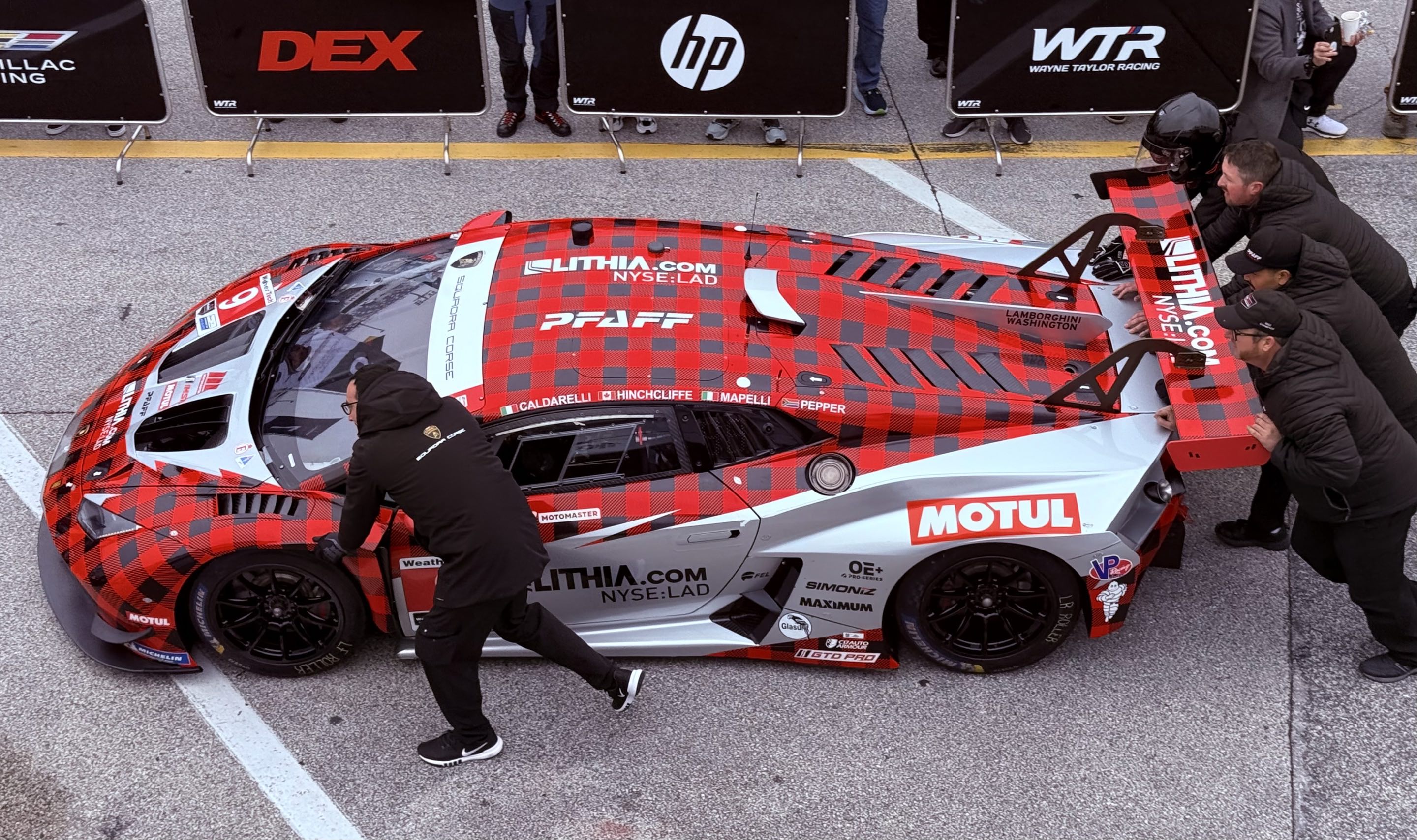 A Lamborghini Huracan GT3 with a plaid livery being pushed through the garage area by crew
